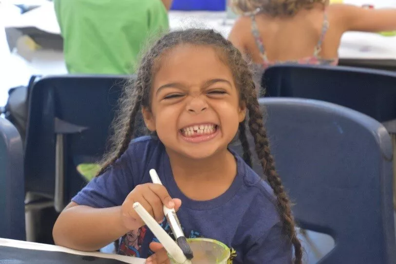 A young child smiles at the viewer as she holds a paintbrush in an art class.