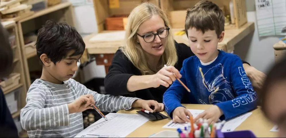 A teacher assists two young students working at a table.