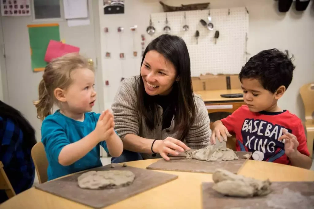 A teacher helps two young students working with clay at a table.