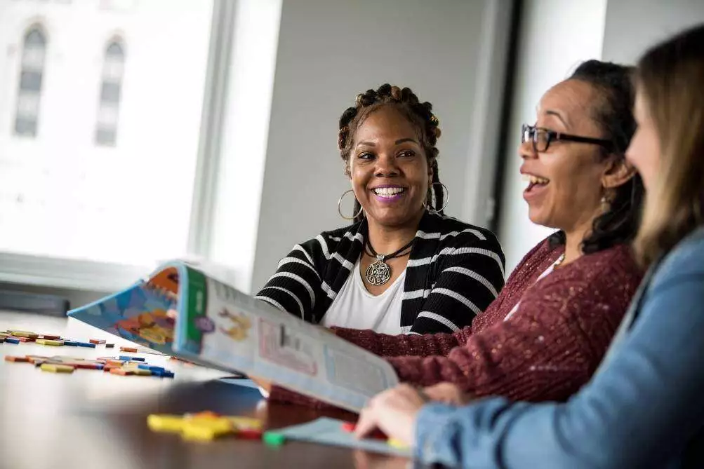 A coach smiles on as two teachers excitedly look through teaching materials.