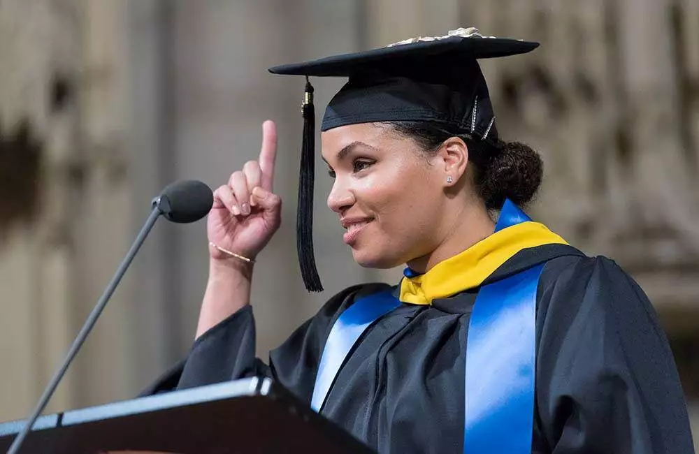 A graduate in a cap and gown stands at a podium holding up one finger in celebration.