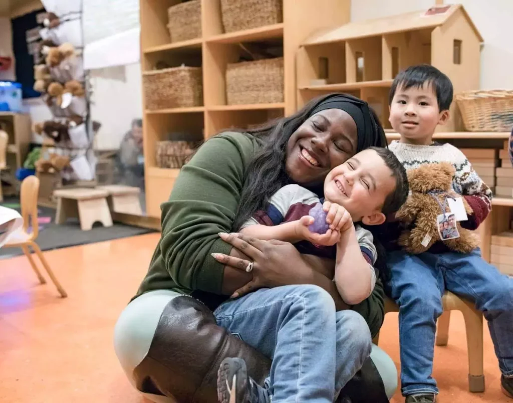 A caring teacher sits on the floor embracing a smiling early elementary student.