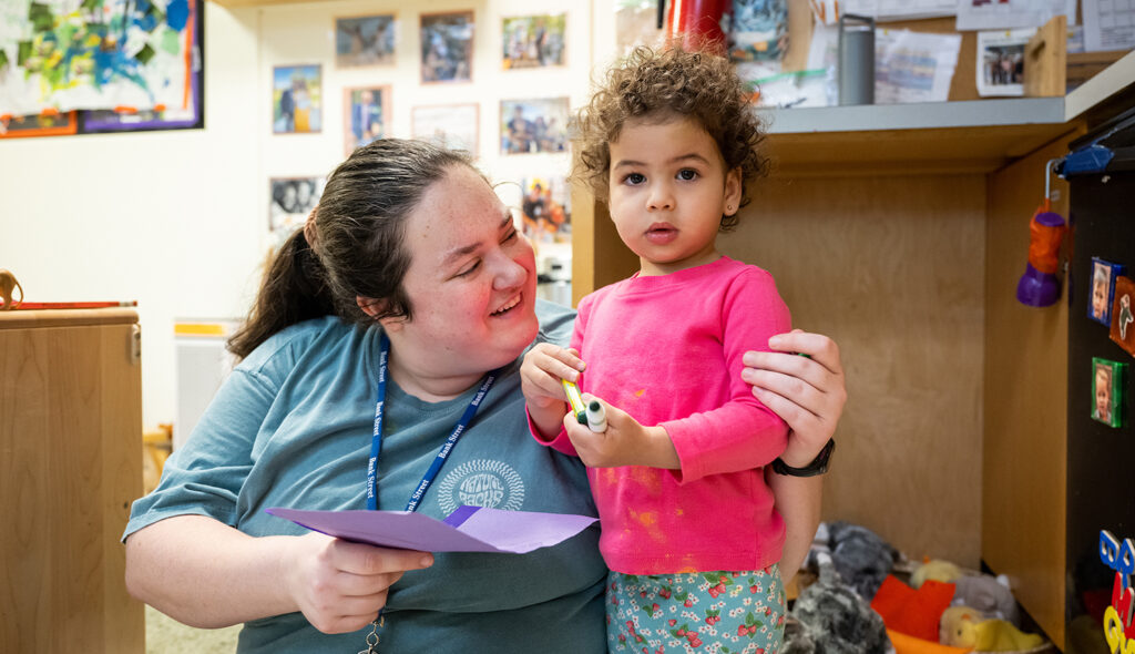 A teacher wearing a lanyard and holding a purple piece of paper smiles at a young student who holds a crayon and a marker.