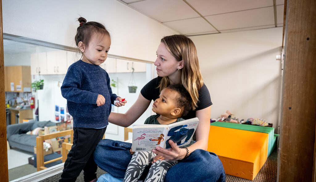 A teacher looks at one young child while reading aloud to another young child seated in her lap.