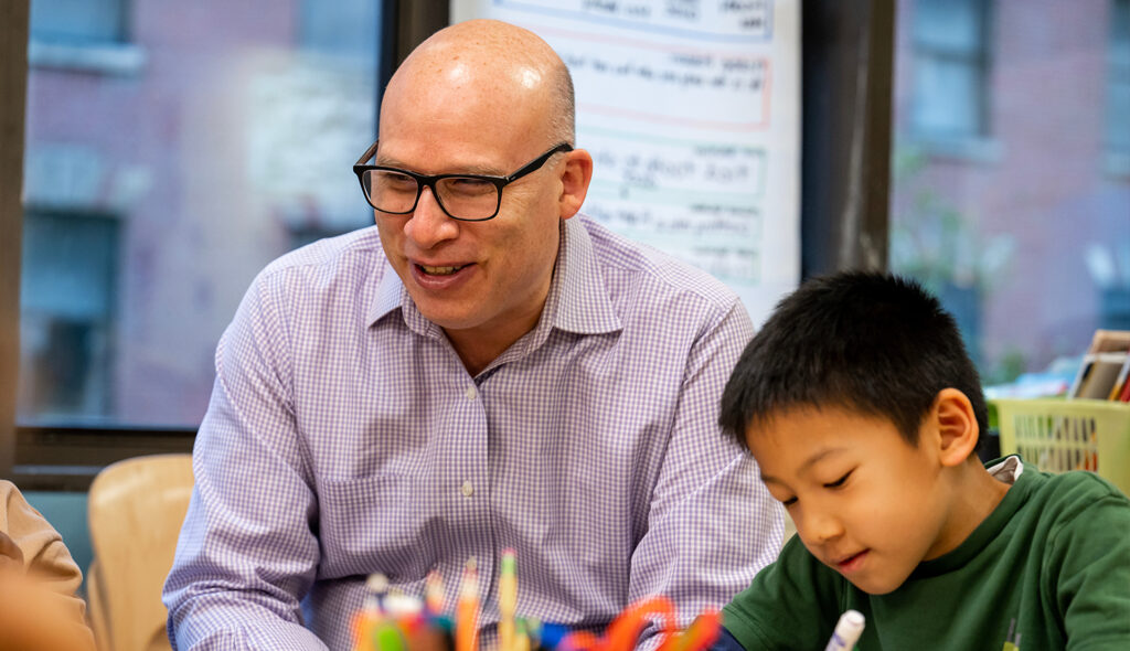 A teacher smiles off-camera as a young student colors with markers.